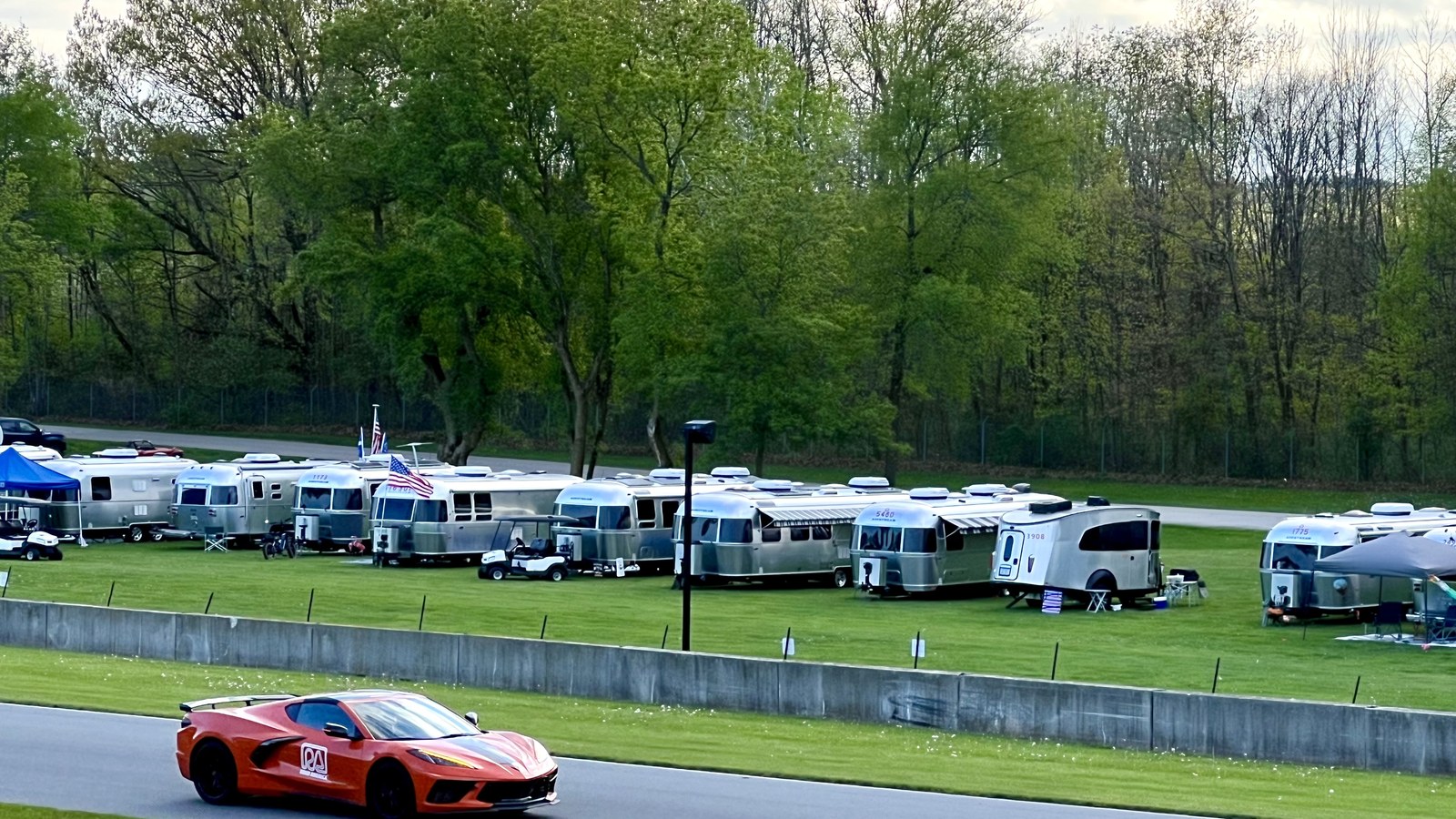 Orange sports car on the Road America track with a row of Airstream trailers on the infield grass, May 2025.