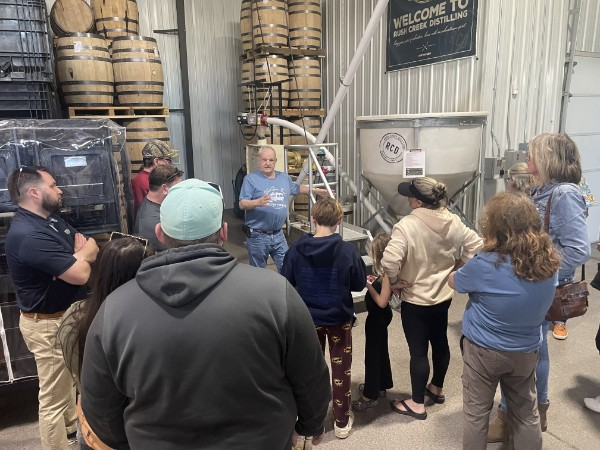Distilling tour guide speaking to guests beside barrels and Rush Creek equipment.